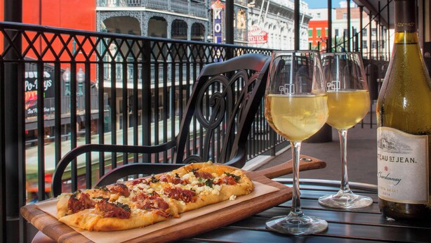 En pizza på en träbricka, två glas vitt vin och en vinflaska på ett bord med utsikt över byggnader i bakgrunden.
