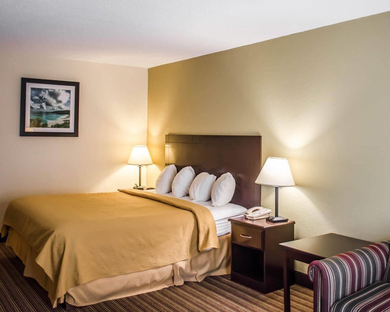 Une chambre d’hôtel avec un lit, des lampes de chevet, un téléphone, une petite table, une chaise et un tableau encadré au mur.