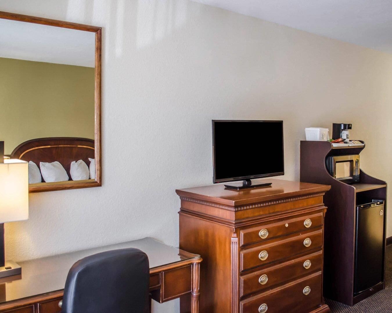 A hotel room with a wooden dresser, a television on top, a desk with a lamp, and a mirror on the wall.