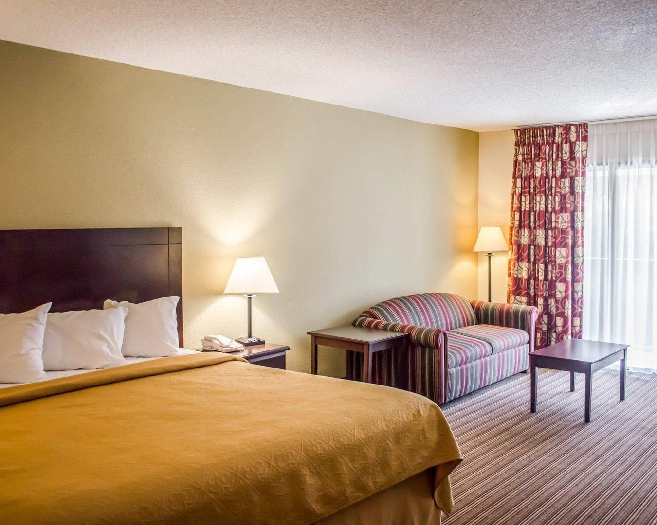 A hotel room with a bed, a sofa, a small table, a lamp, and a window with curtains.