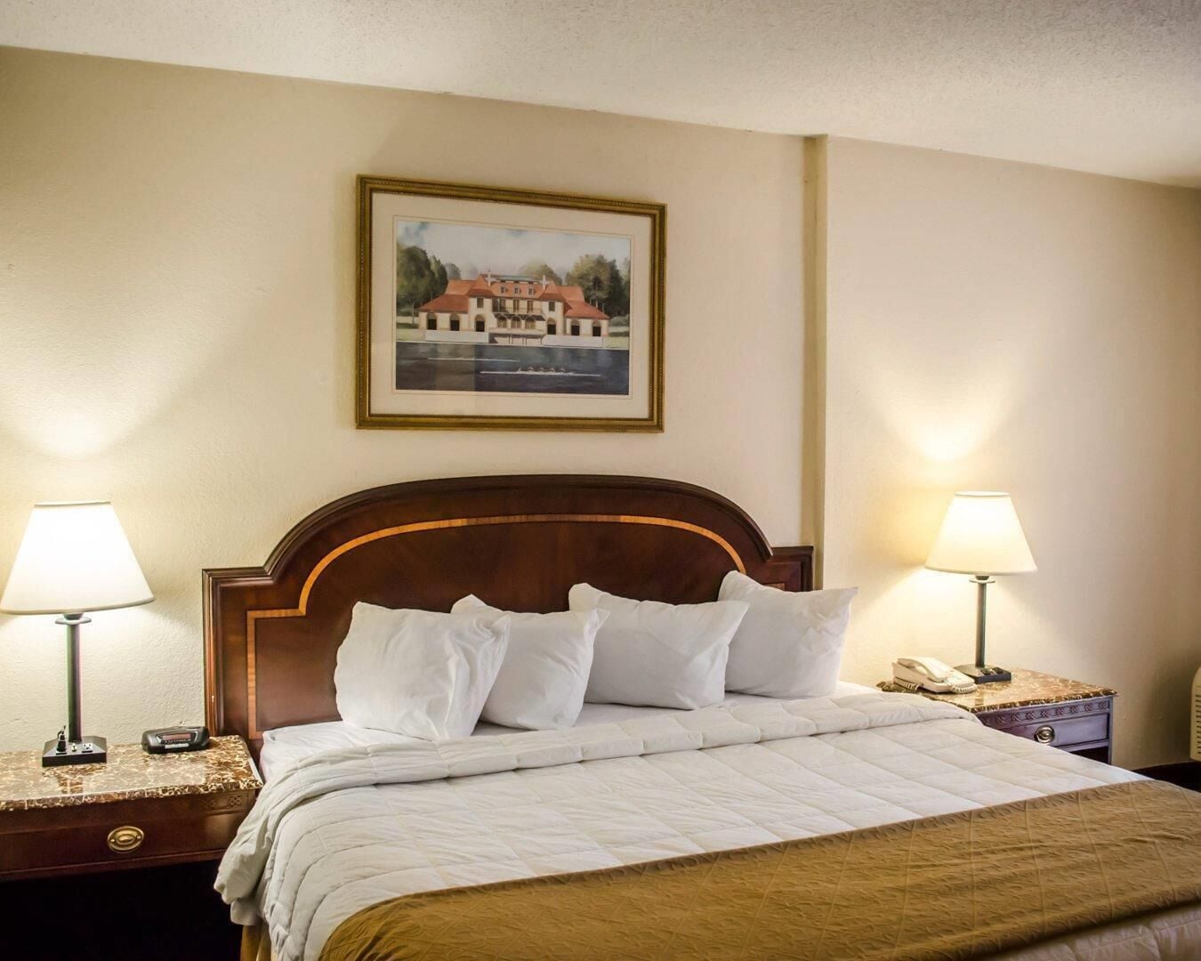 A hotel room with a bed, two bedside lamps, a nightstand with a phone, and a framed picture on the wall.
