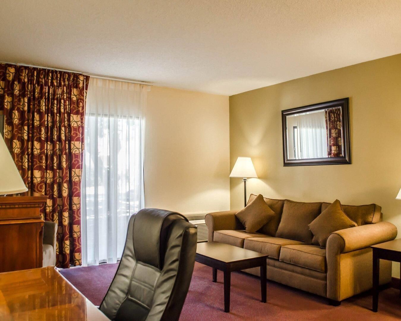 A hotel room with a leather chair, a sofa, a coffee table, a lamp, and a large window with curtains.