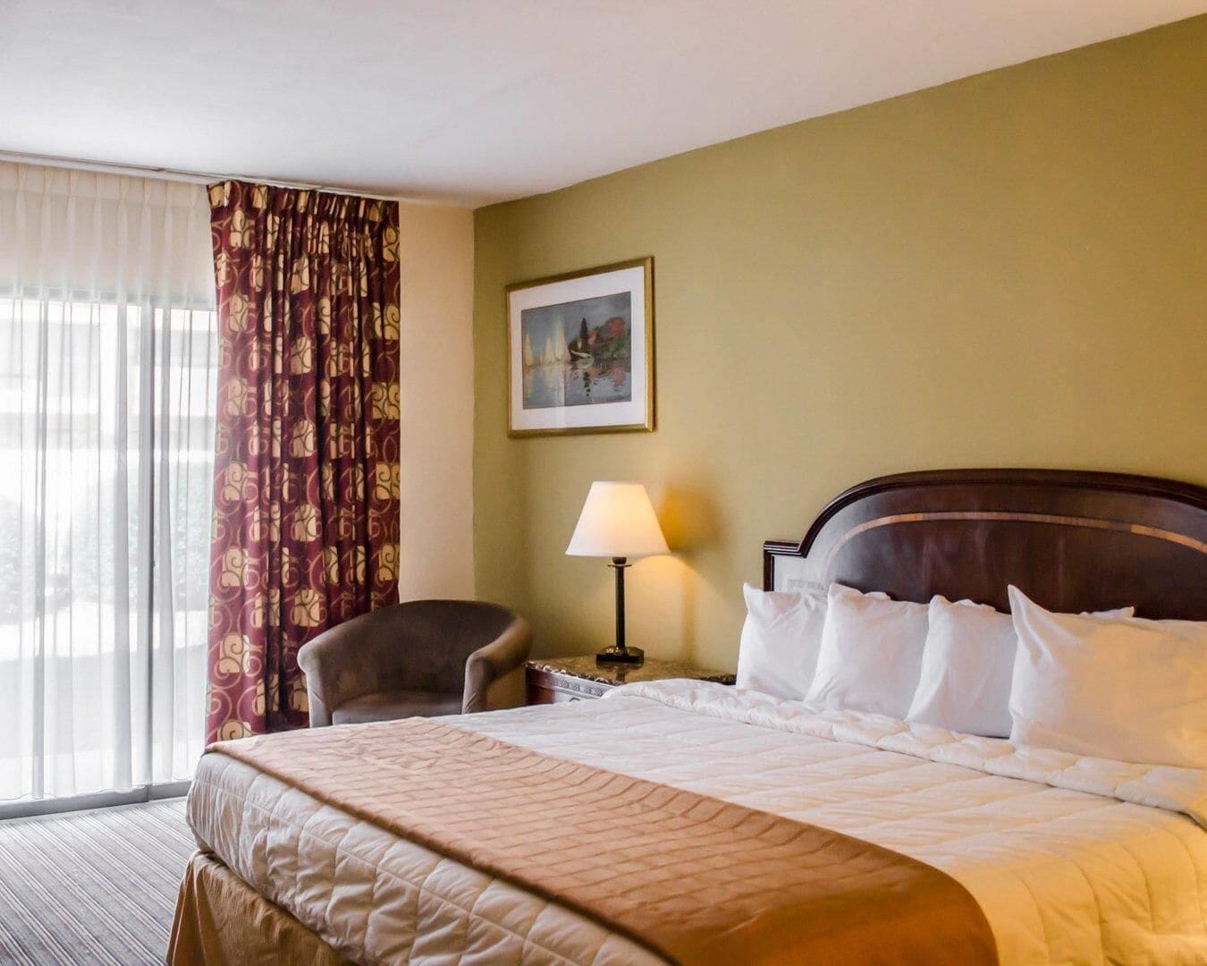 A hotel room with a bed, a chair, a nightstand with a lamp, a painting on the wall, and a window with curtains.