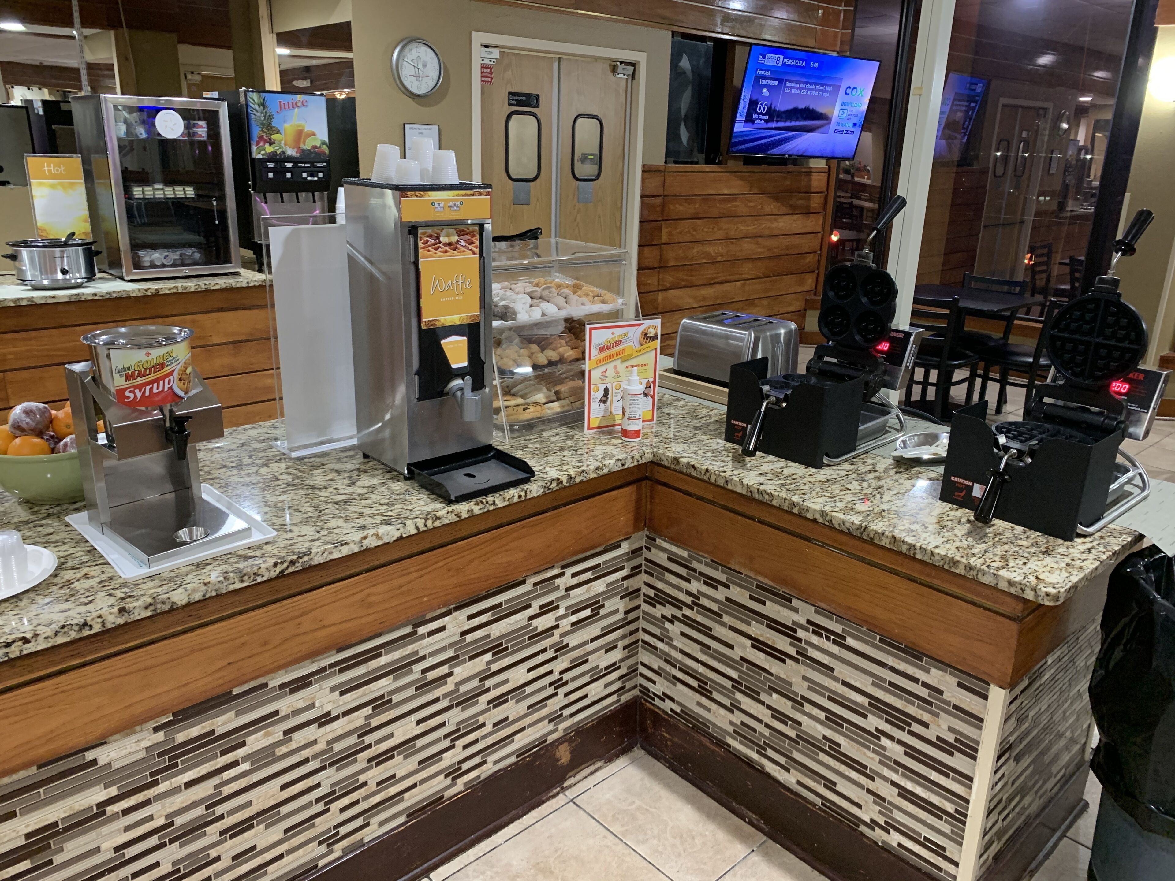 Una estación de desayuno autoservicio con máquinas para hacer gofres, una cafetera y un dispensador de sirope.