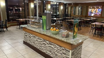 A hotel lobby with a central counter featuring a fruit bowl, a coffee machine, and a fresh juice dispenser.
