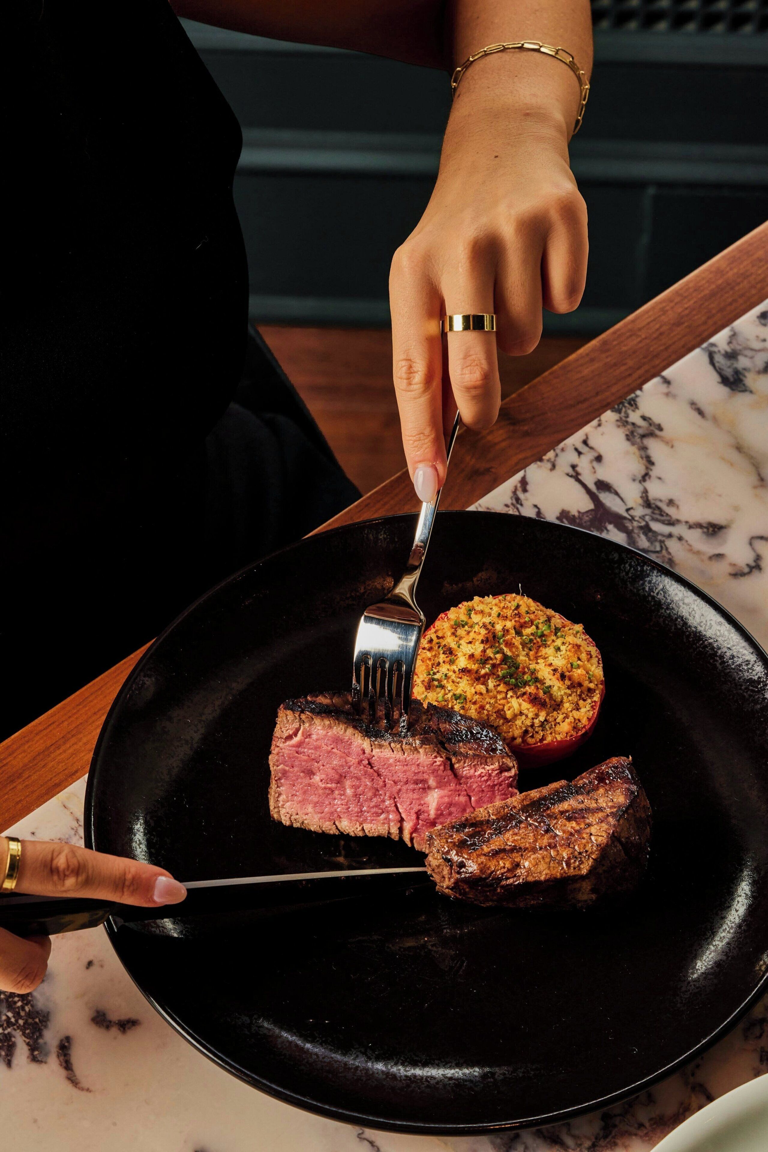 A person cutting a steak on a black plate with a fork and knife.