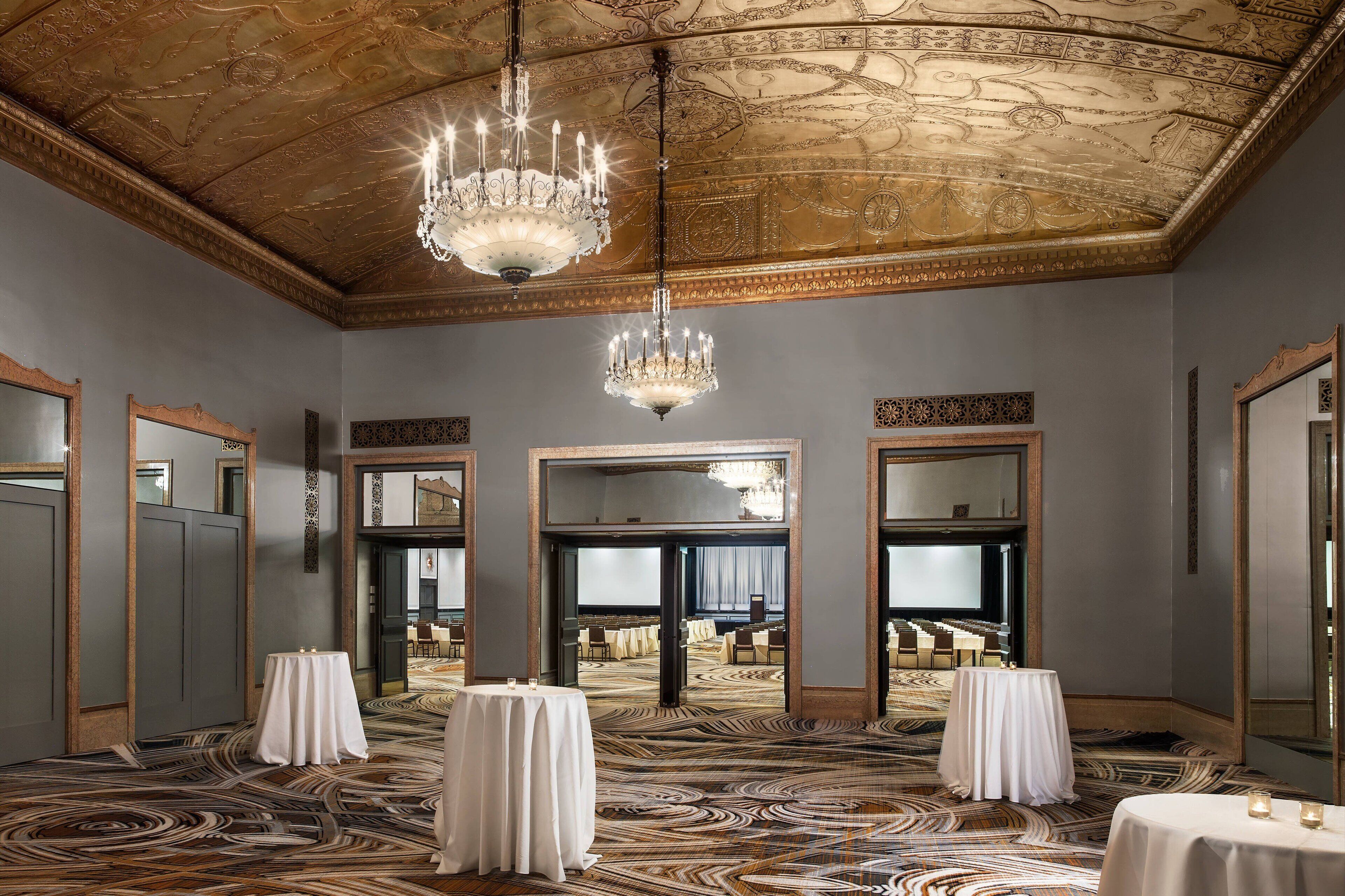 A grand room with ornate ceiling and chandeliers, large mirrors, and a patterned carpet.
