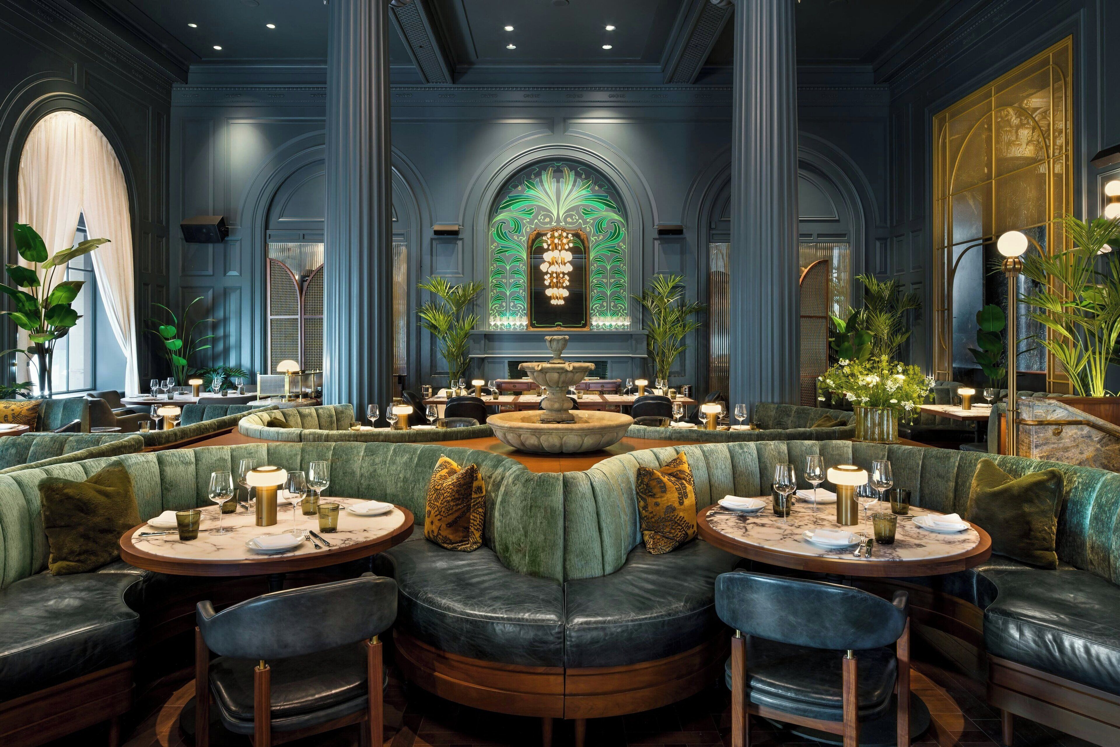 Uma sala de jantar luxuosa com uma fonte central, assentos estofados em verde e várias mesas de jantar.