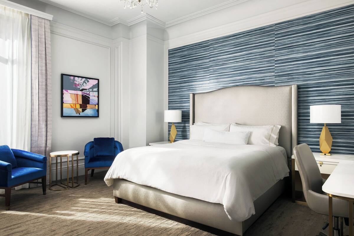 Een hotelkamer met een groot bed, twee blauwe fauteuils, een klein rond tafeltje en een bureau met een lamp.