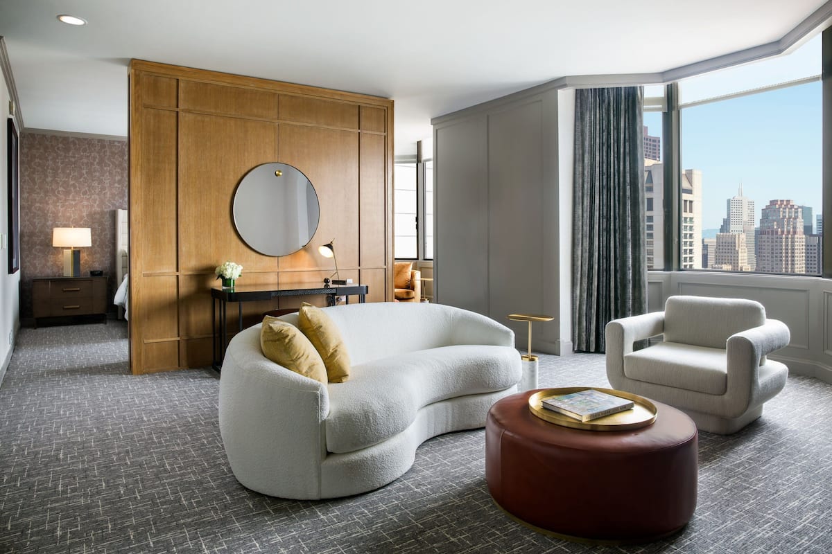 Een moderne hotelkamer met een gebogen bank, een rond poefje, een stoel, een grote spiegel en uitzicht op de stad door een groot raam.