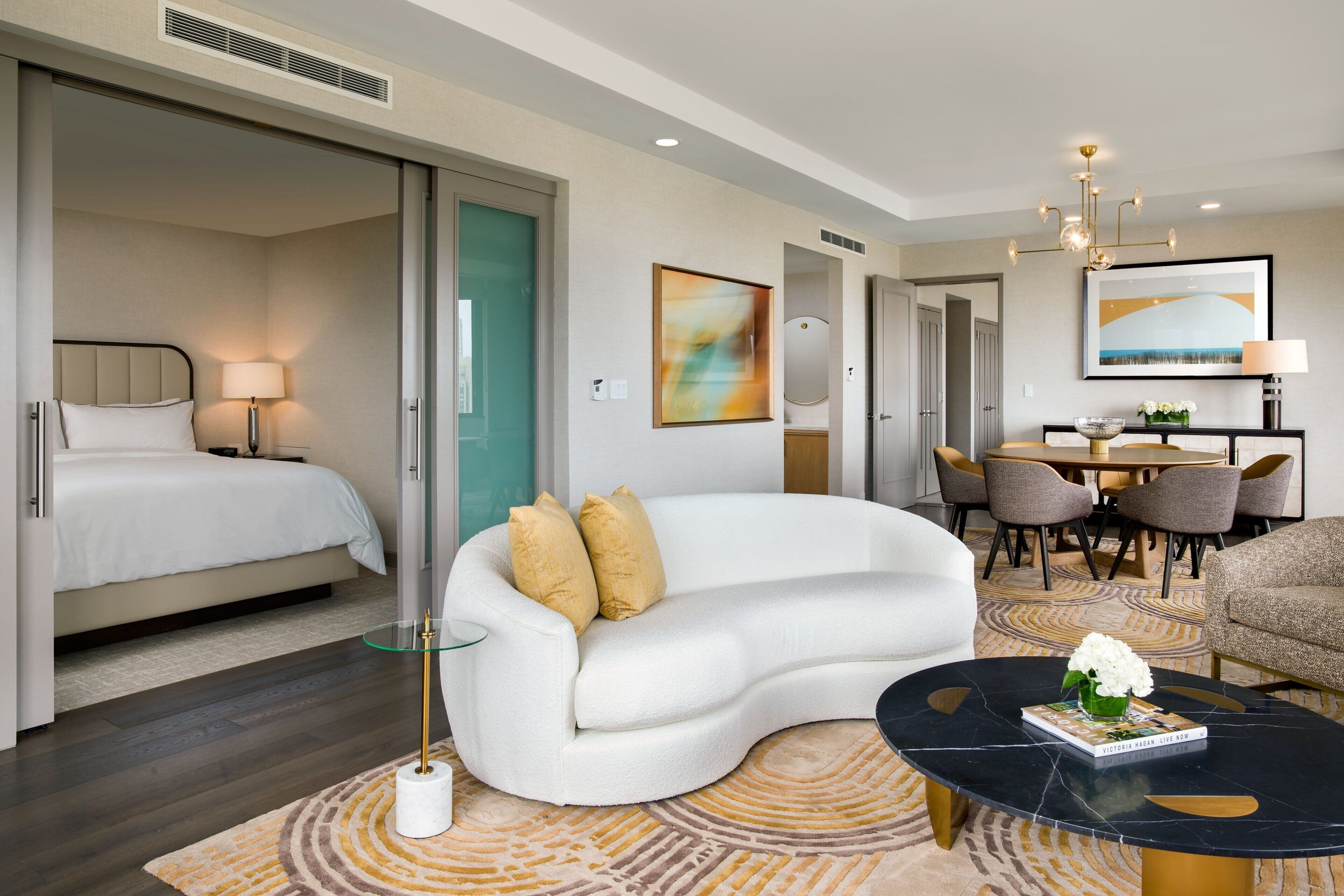 Ein modernes Hotelzimmer mit einer gebogenen weißen Couch, einem Glastisch, einem Bett mit weißer Bettwäsche und einem Essbereich mit Tisch und Stühlen.