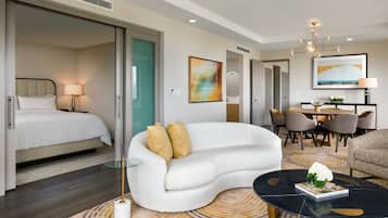 Ein modernes Hotelzimmer mit einer gebogenen weißen Couch, einem Glastisch, einem Bett mit weißer Bettwäsche und einem Essbereich mit Tisch und Stühlen.