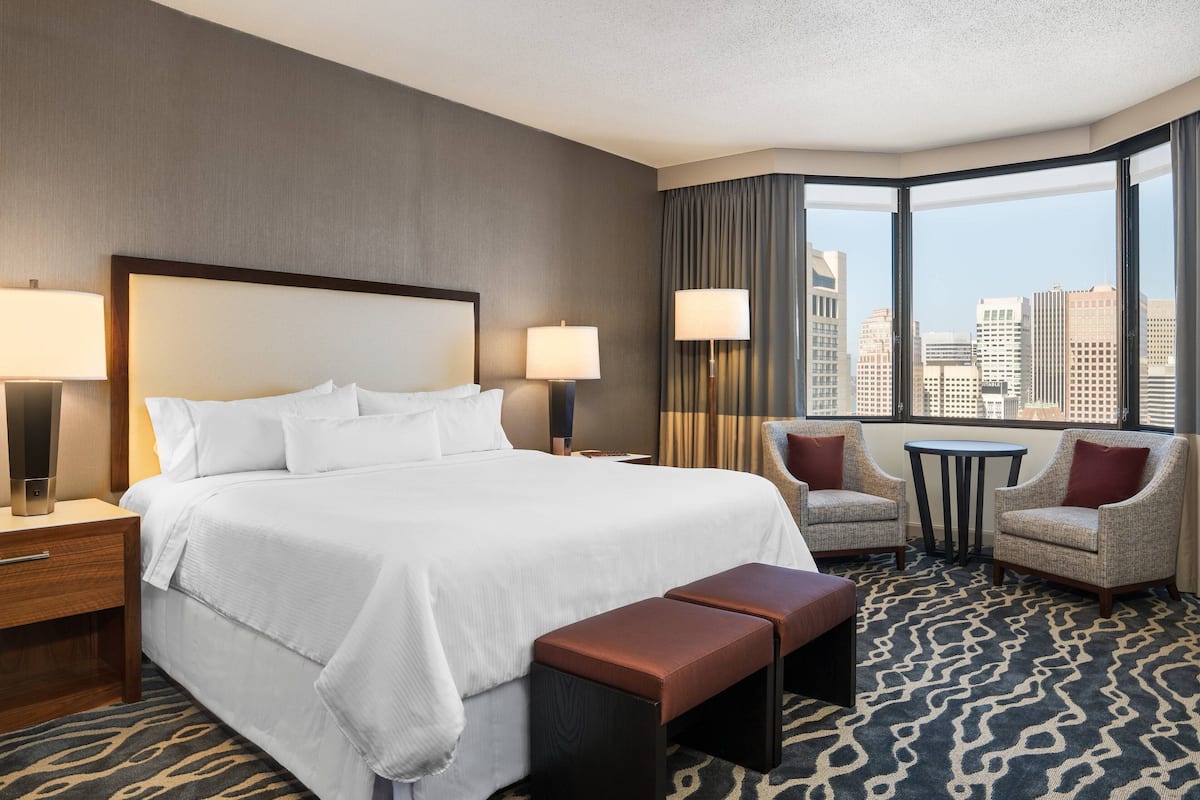 Een hotelkamer met een groot bed, twee nachtkastjes, een stoel, een kleine tafel en uitzicht op de stad door grote ramen.
