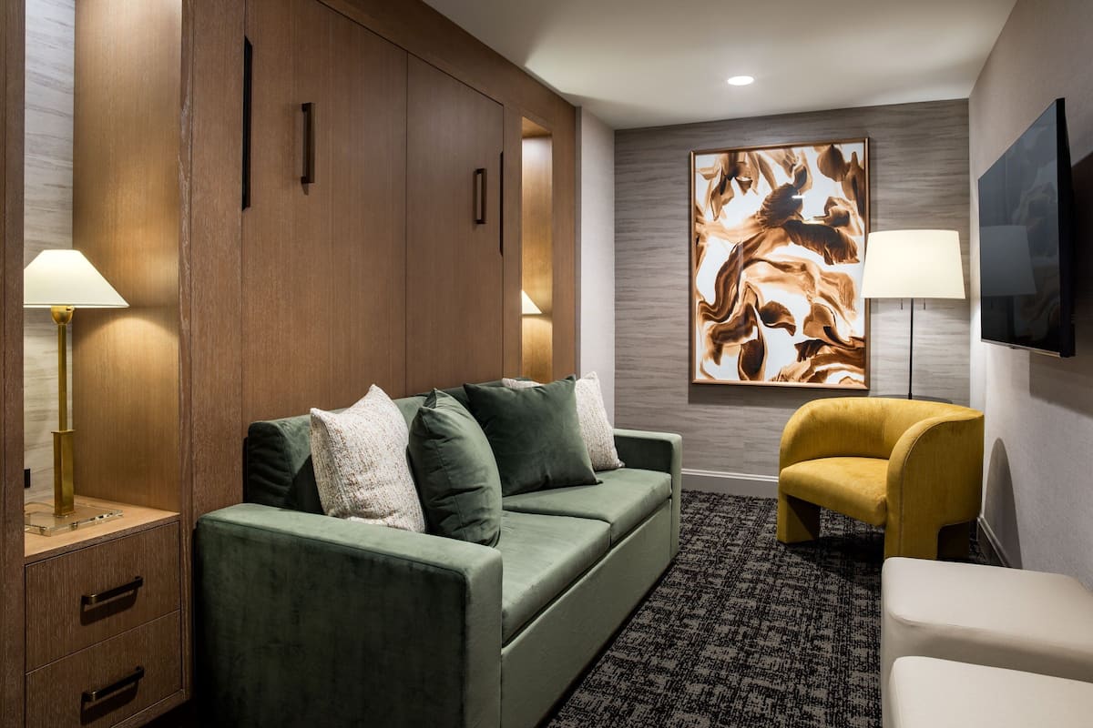 Hotelkamer met een groene bank, een gele fauteuil, een flatscreen televisie en een groot abstract schilderij aan de muur.
