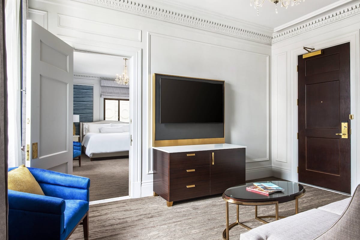 Een hotelkamer met een grote flatscreentelevisie, een donker houten kast met gouden handgrepen, een blauwe fauteuil, een ronde glazen salontafel en een grote donkerhouten deur.