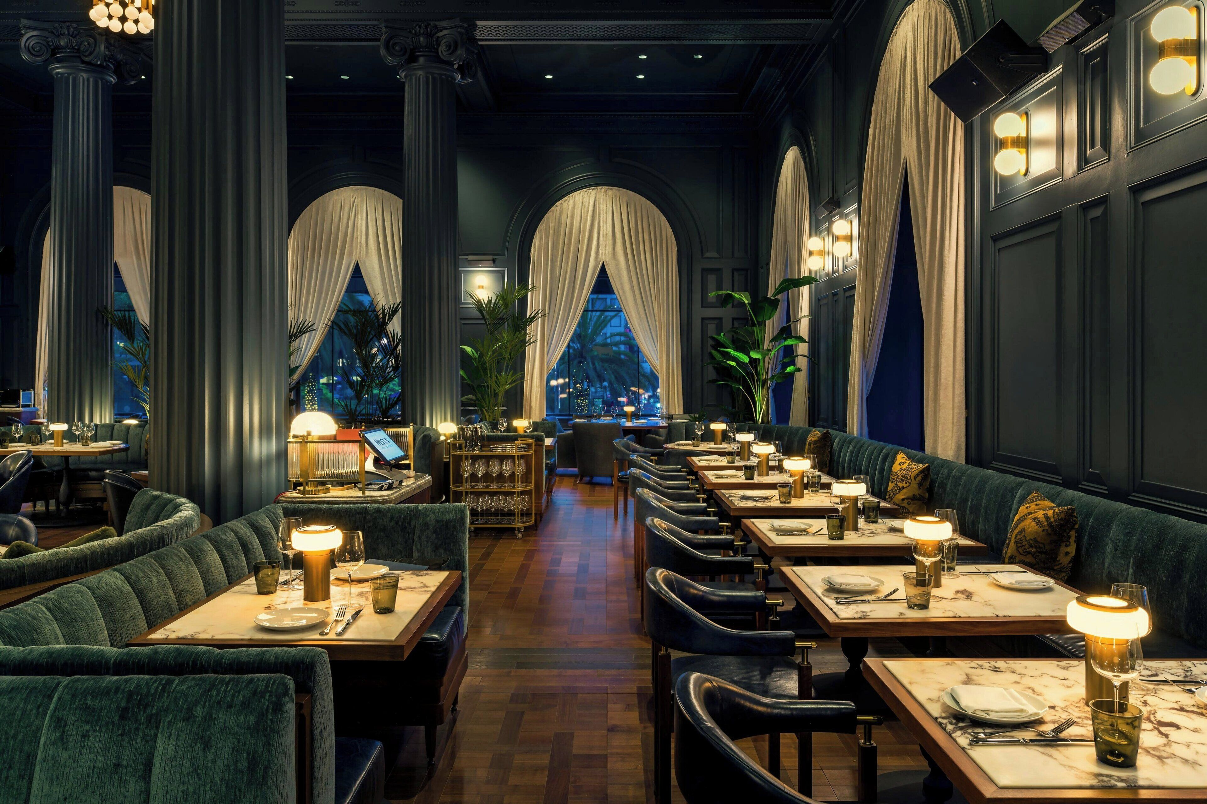 A restaurant with high ceilings, large windows with draped curtains, and a long central aisle leading to the back. Tables are set with candles and tableware.