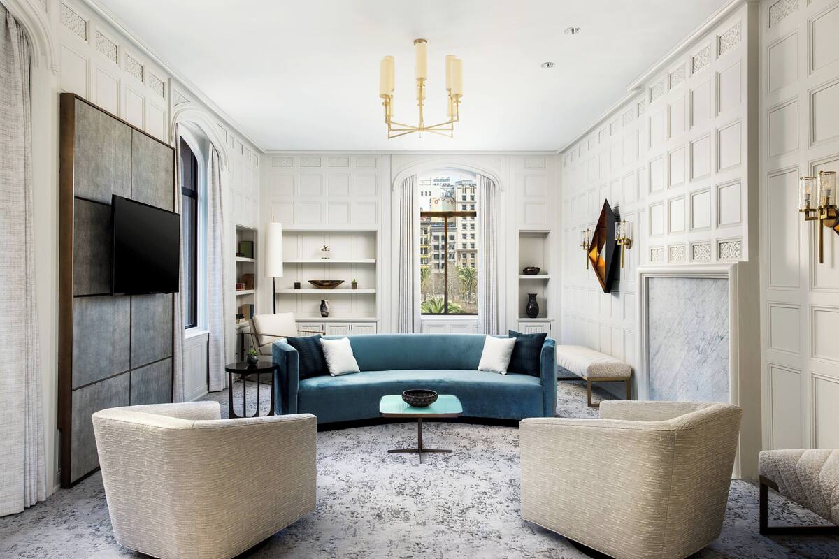 Een moderne woonkamer met een blauwe bank, beige fauteuils, een salontafel, een flatscreen-tv en een kroonluchter.