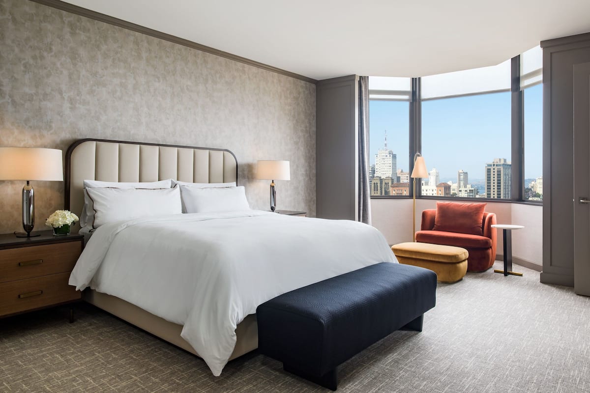 Een moderne hotelkamer met een groot bed, twee bedlampen, een bankje, een rode stoel en uitzicht op de stad door grote ramen.