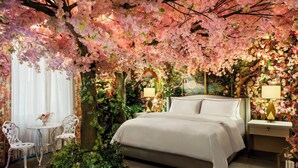 Une chambre à coucher avec un grand lit, deux tables de chevet, une petite table ronde avec des chaises et une grande fenêtre ornée de rideaux légers.