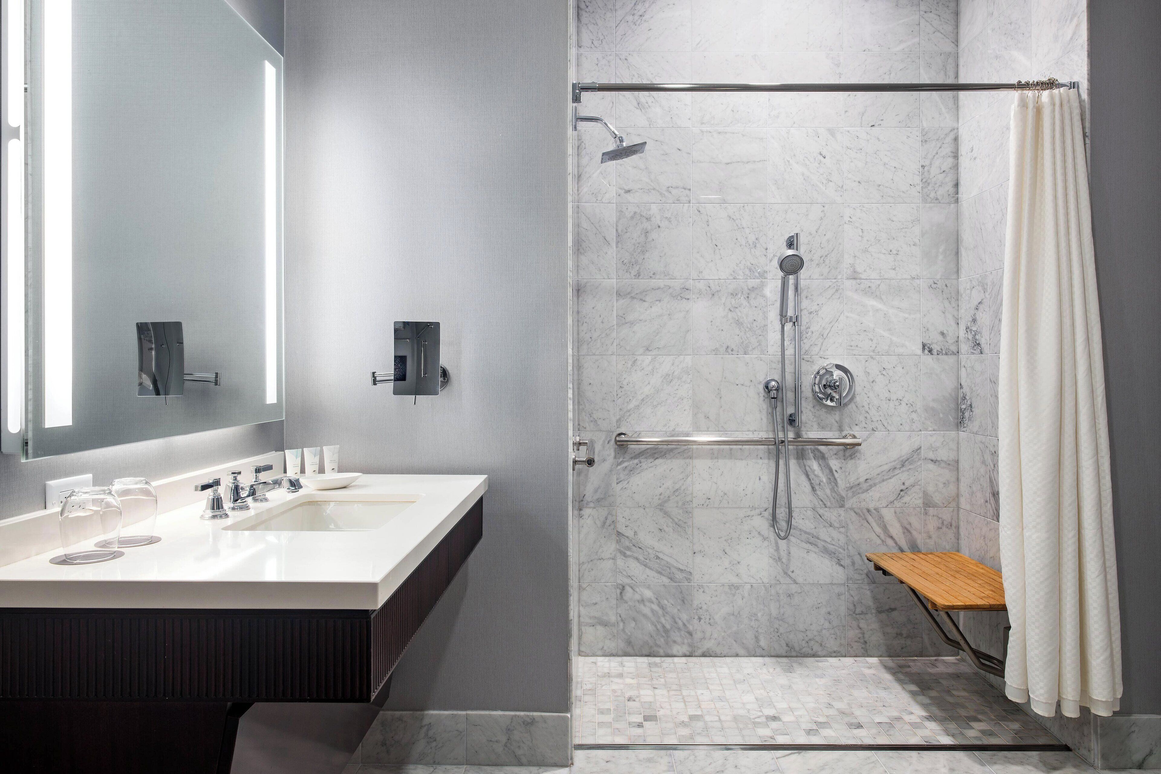 A modern bathroom with a large mirror, two sinks, a shower with a glass door, and a wooden bench.