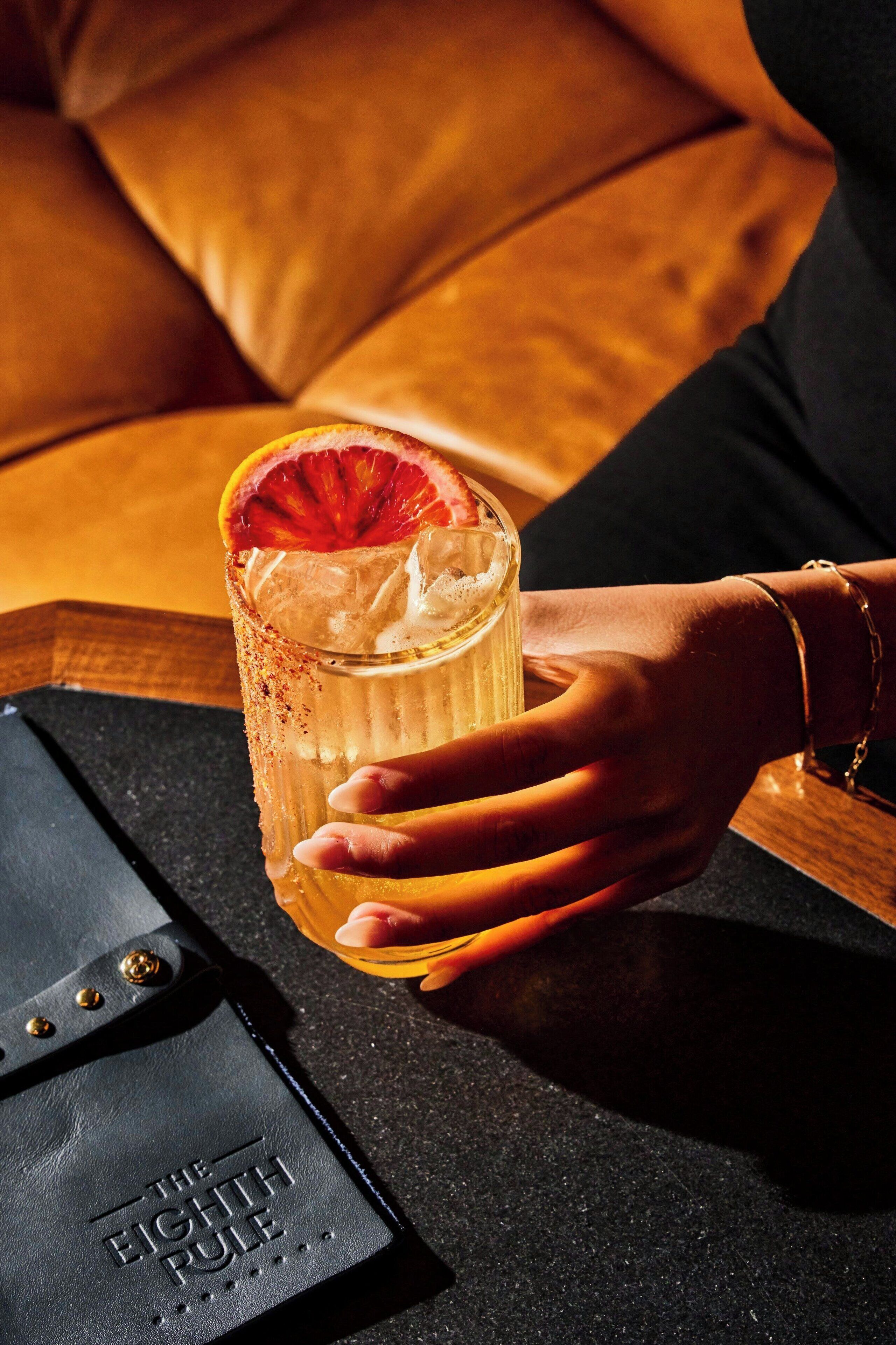 A hand holding a cocktail glass with a slice of fruit on the rim, placed on a table next to a black leather wallet with embossed text.