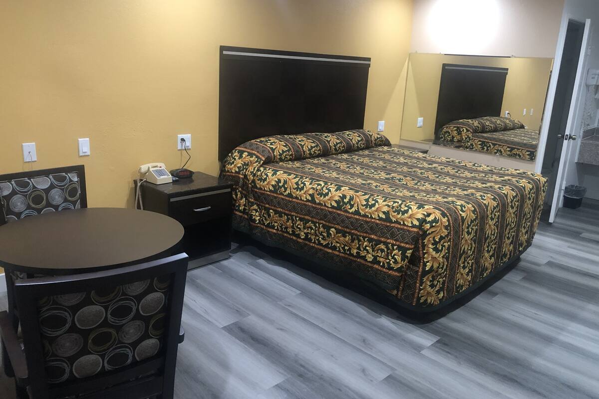 Une chambre d’hôtel avec un lit, un bureau et une chaise. Il y a un miroir sur le mur et une porte qui donne sur une autre pièce.