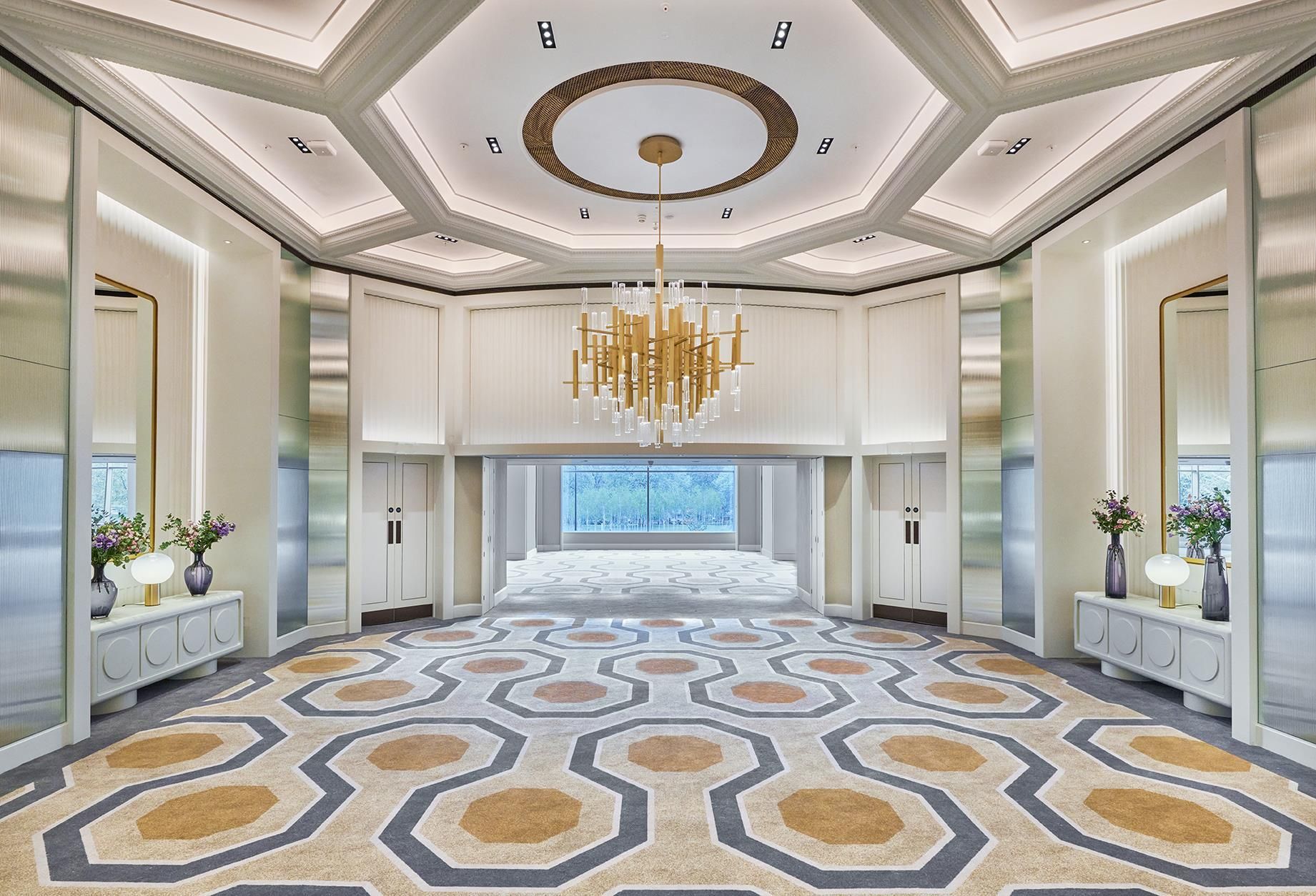A spacious hotel lobby with a patterned carpet, a large chandelier, and a view through the glass doors.