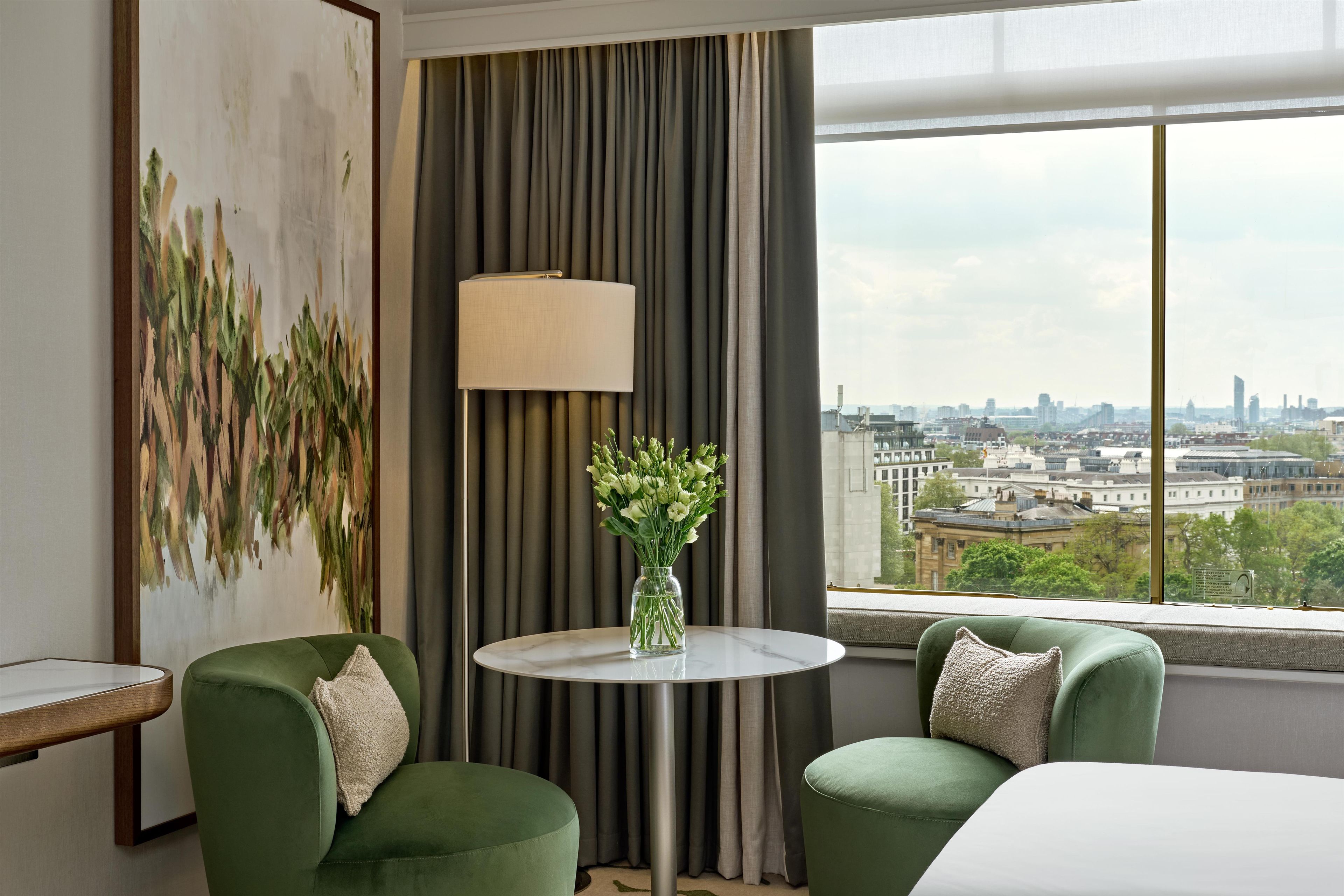 A hotel room with a city view, two green armchairs, a round table with a vase of flowers, and a large painting on the wall.