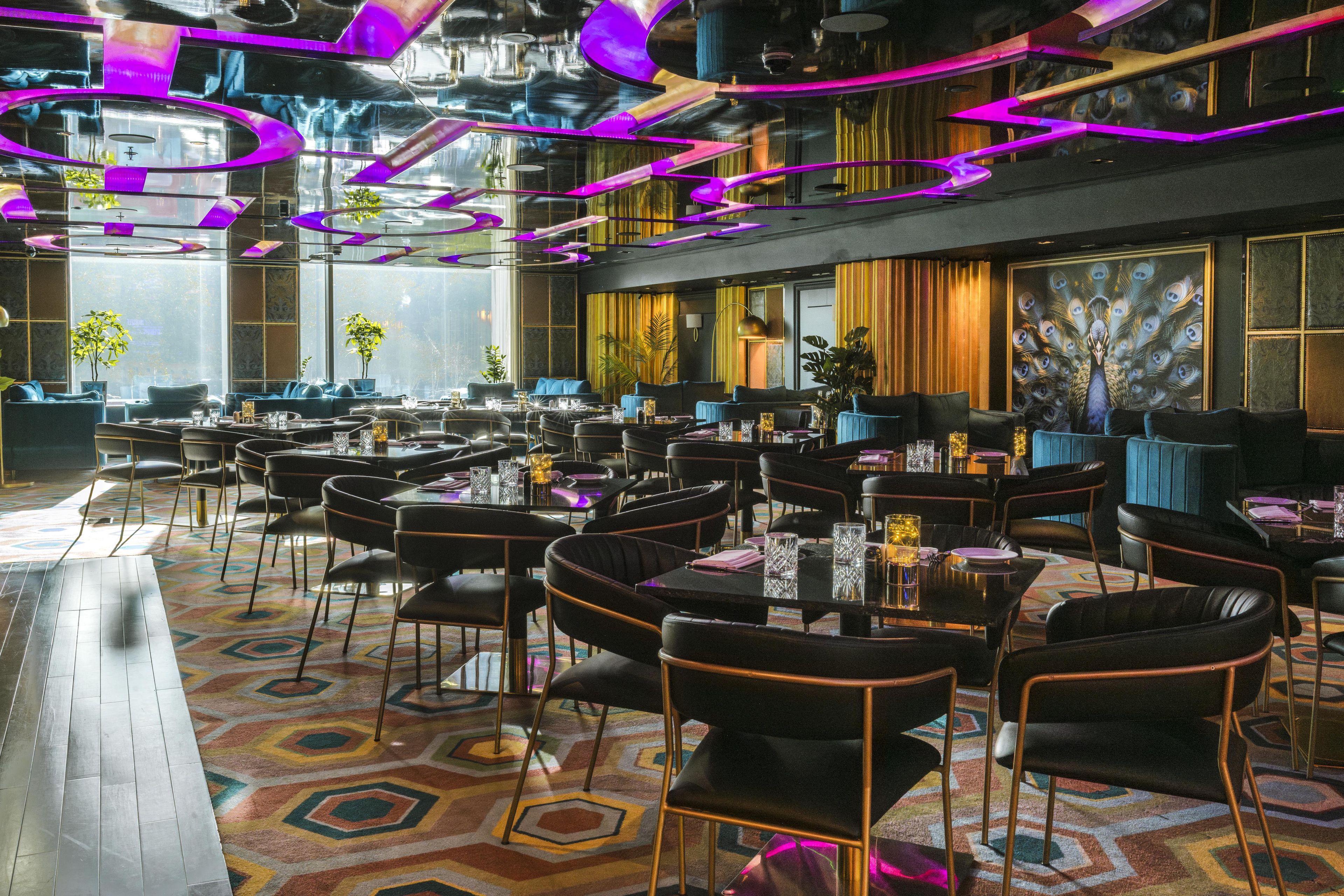 A spacious restaurant with multiple dining tables and chairs, large windows, and a colorful geometric carpet.