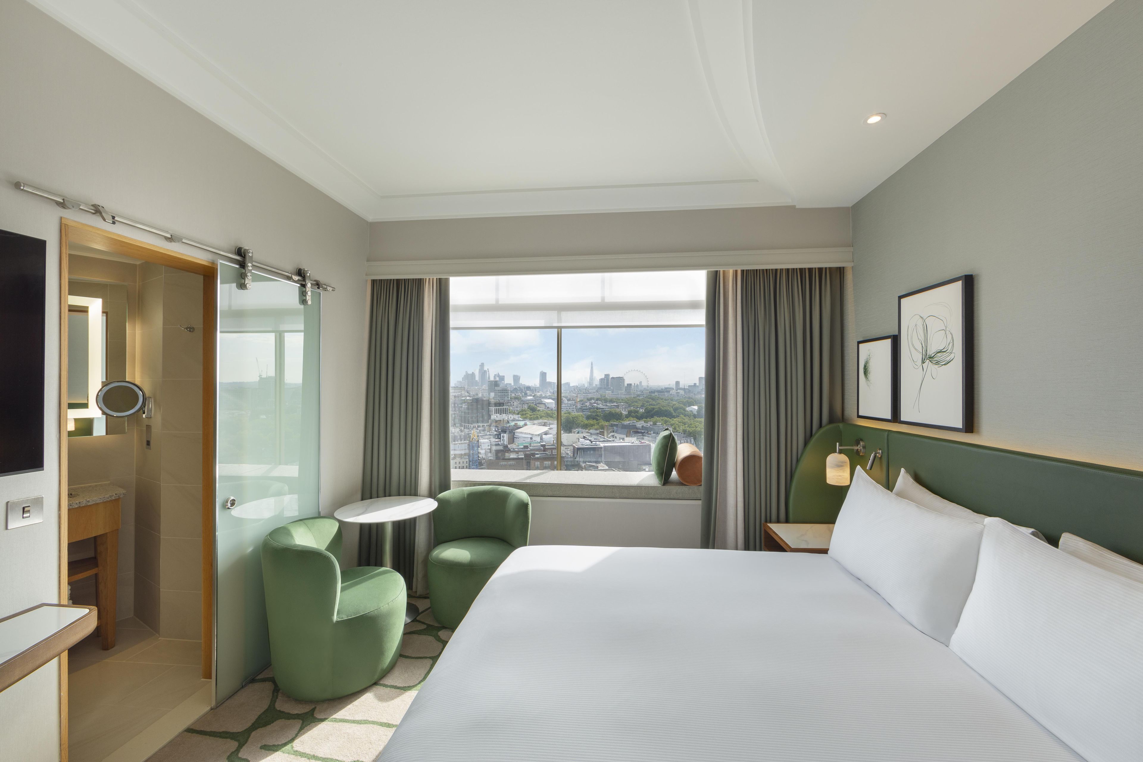 A hotel room with a bed, a small table, two chairs, a TV, and a window with a city view.