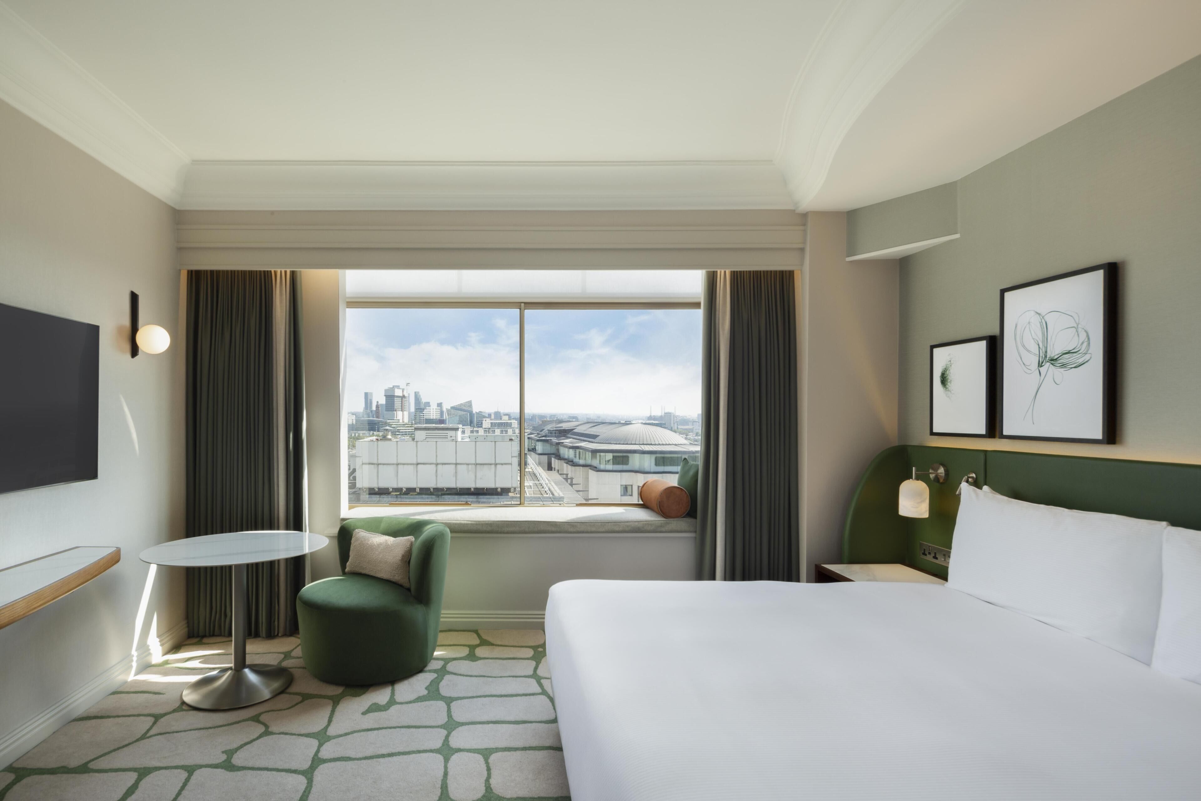 A hotel room with a bed, a small round table, a green armchair, a window with a city view, and two framed artworks on the wall.