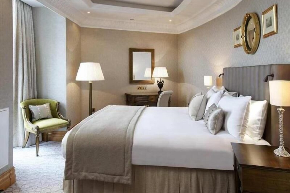 A hotel room with a large bed, two bedside lamps, a green armchair, a dresser, and a mirror on the wall.