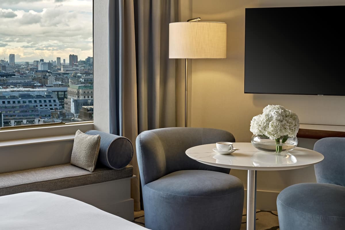 Ett hotellrum med utsikt över staden, en soffa, ett bord med en vas med blommor, en lampa och en tv.