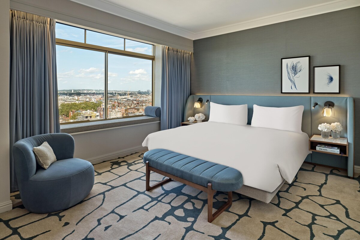 Habitación de hotel con una cama grande, un banco, una silla, una mesita de noche con lámpara y vistas a la ciudad.