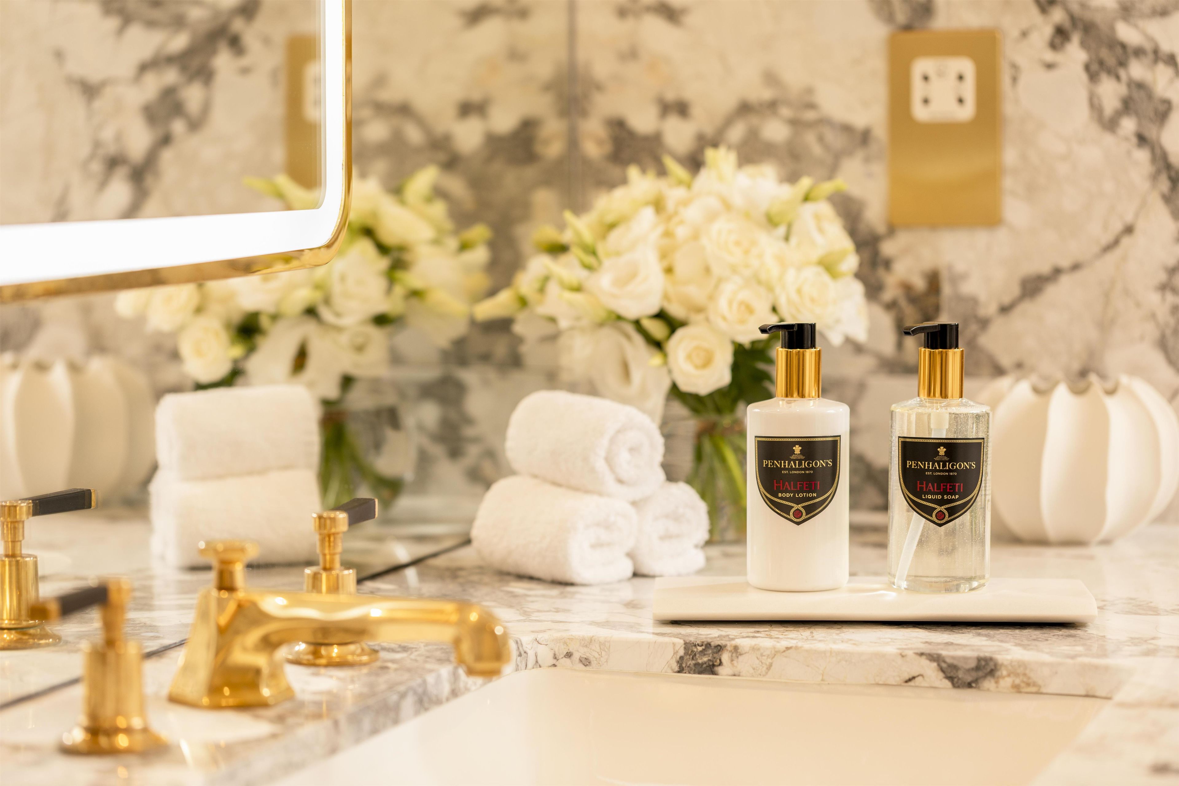 A bathroom vanity with a marble countertop, gold faucets, and two bottles of Penhaligon's Halfeti body lotion and liquid soap.