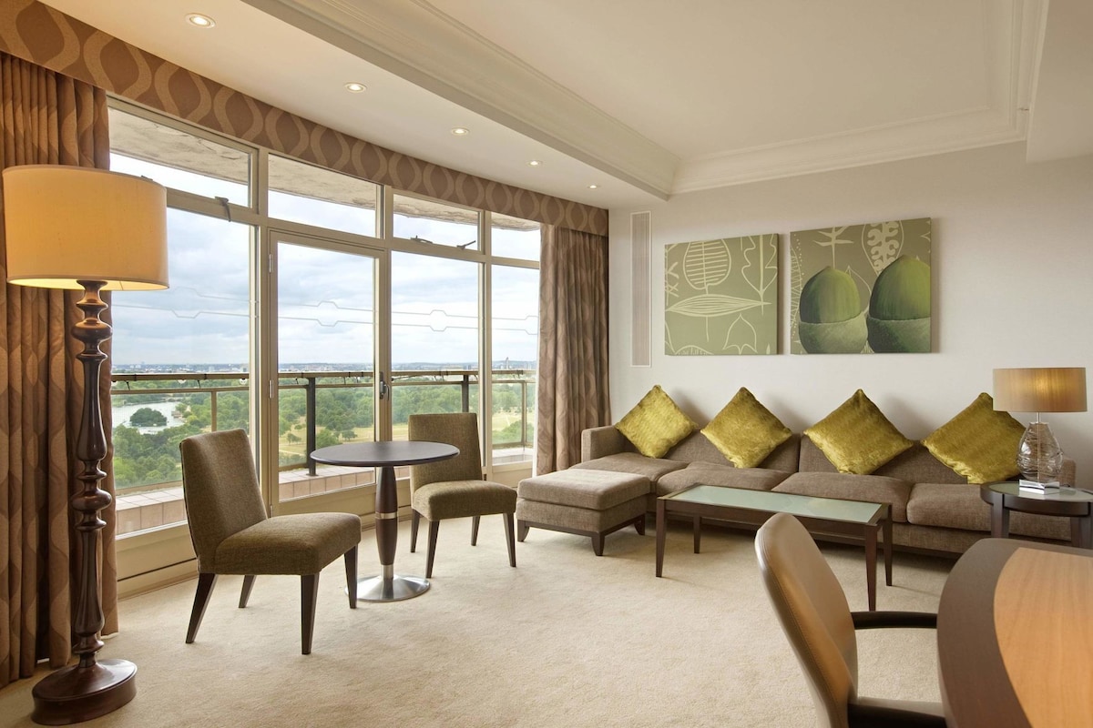 A hotel room with a balcony, a sofa set, a dining table, and a view of a cityscape.