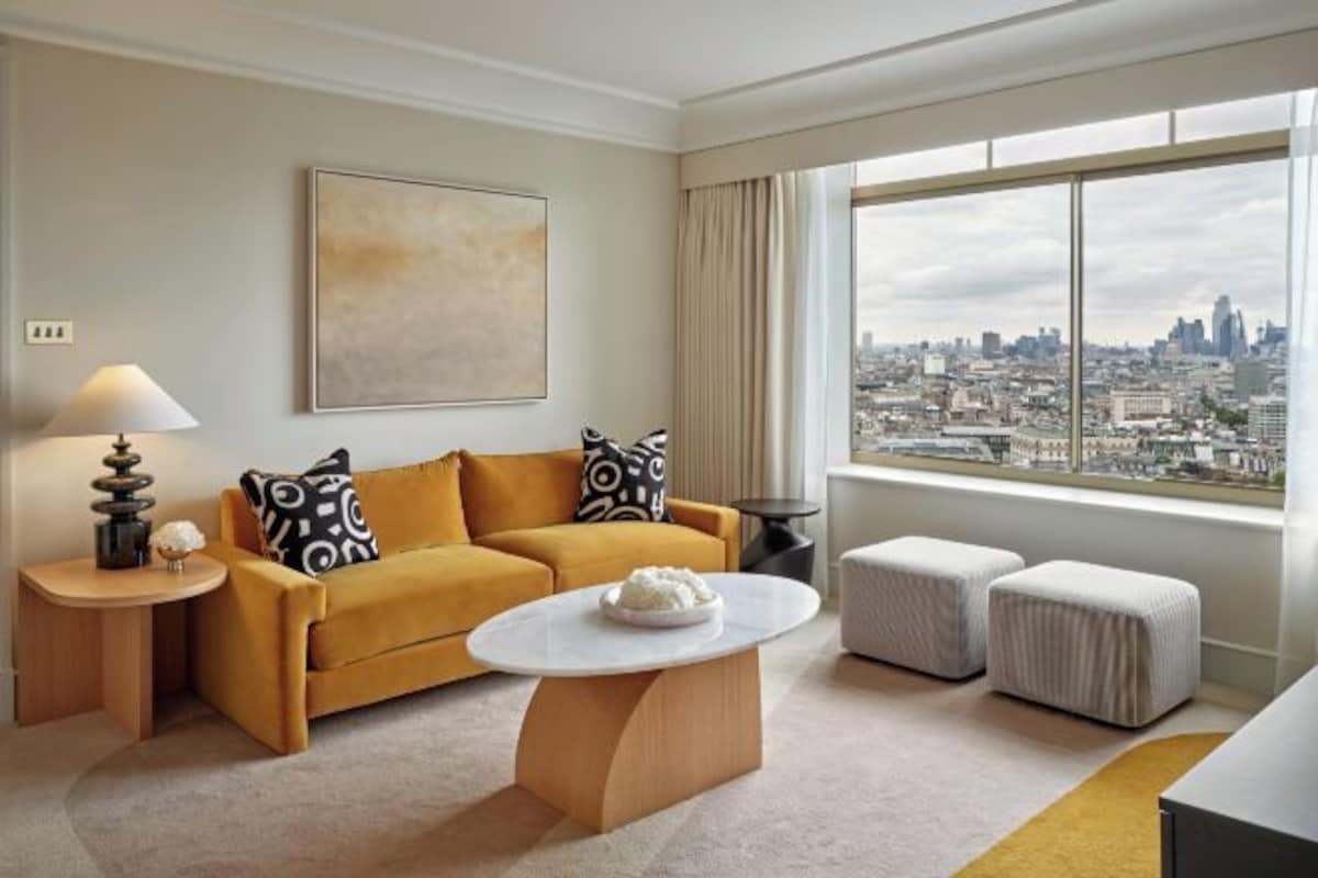 A modern hotel room with a city view, a sofa, ottomans, a round coffee table, and a lamp.