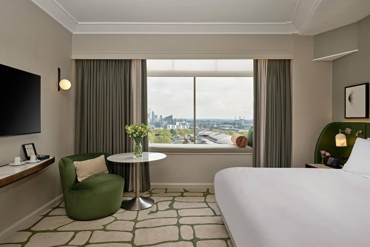 Ett hotellrum med en säng, en grön fåtölj, ett litet bord med en vas, en tv som är monterad på väggen och ett fönster med utsikt över staden.