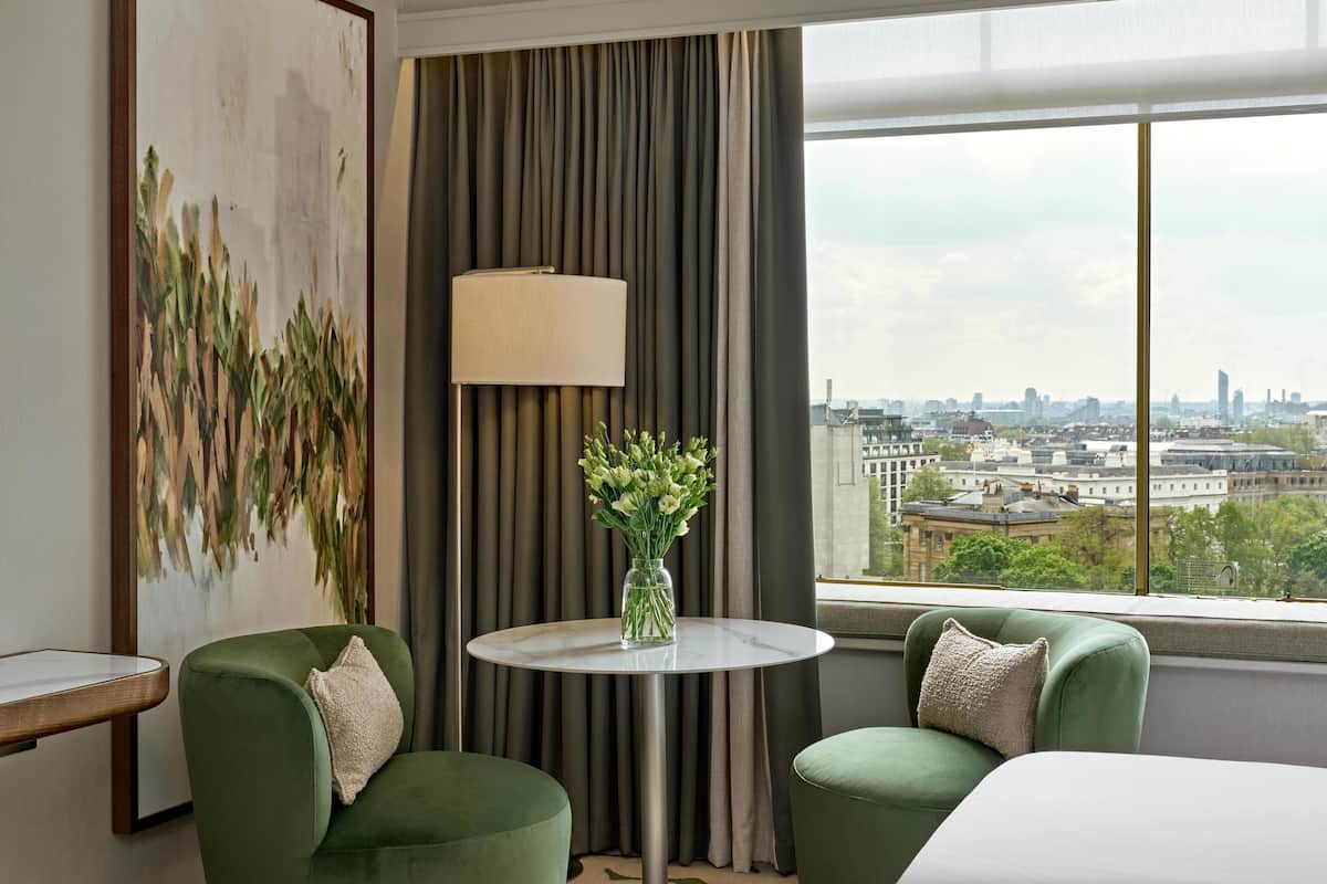 Ett hotellrum med utsikt över staden, ett runt bord med en vas med blommor och två gröna fåtöljer.