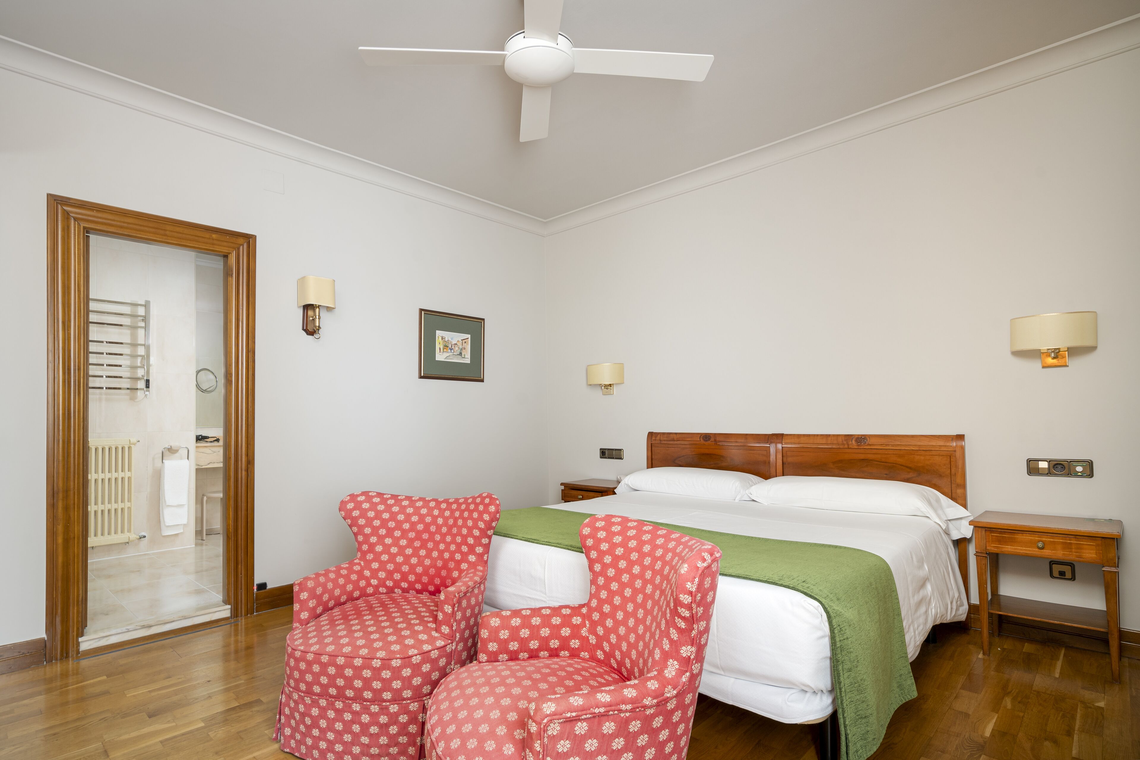 A bedroom with a bed, two nightstands, a wooden chair, a ceiling fan, and a door leading to a bathroom.