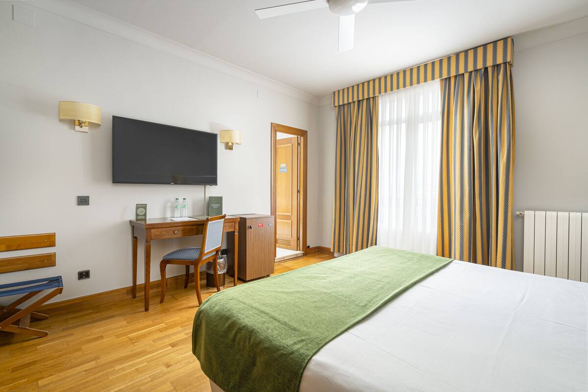 A hotel room with a bed, a desk, a chair, a television, and a window with curtains.