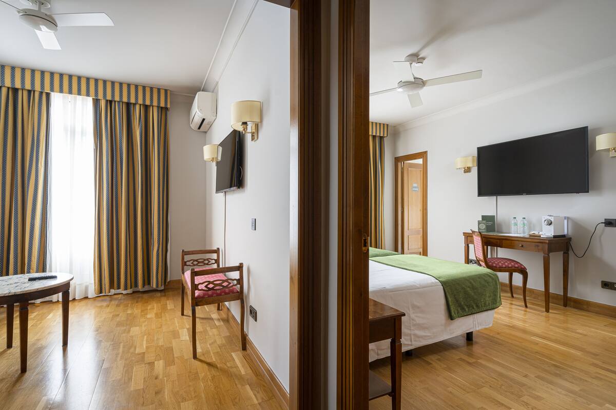 A hotel room with a bed, a desk, a chair, a table, a TV, and a ceiling fan.