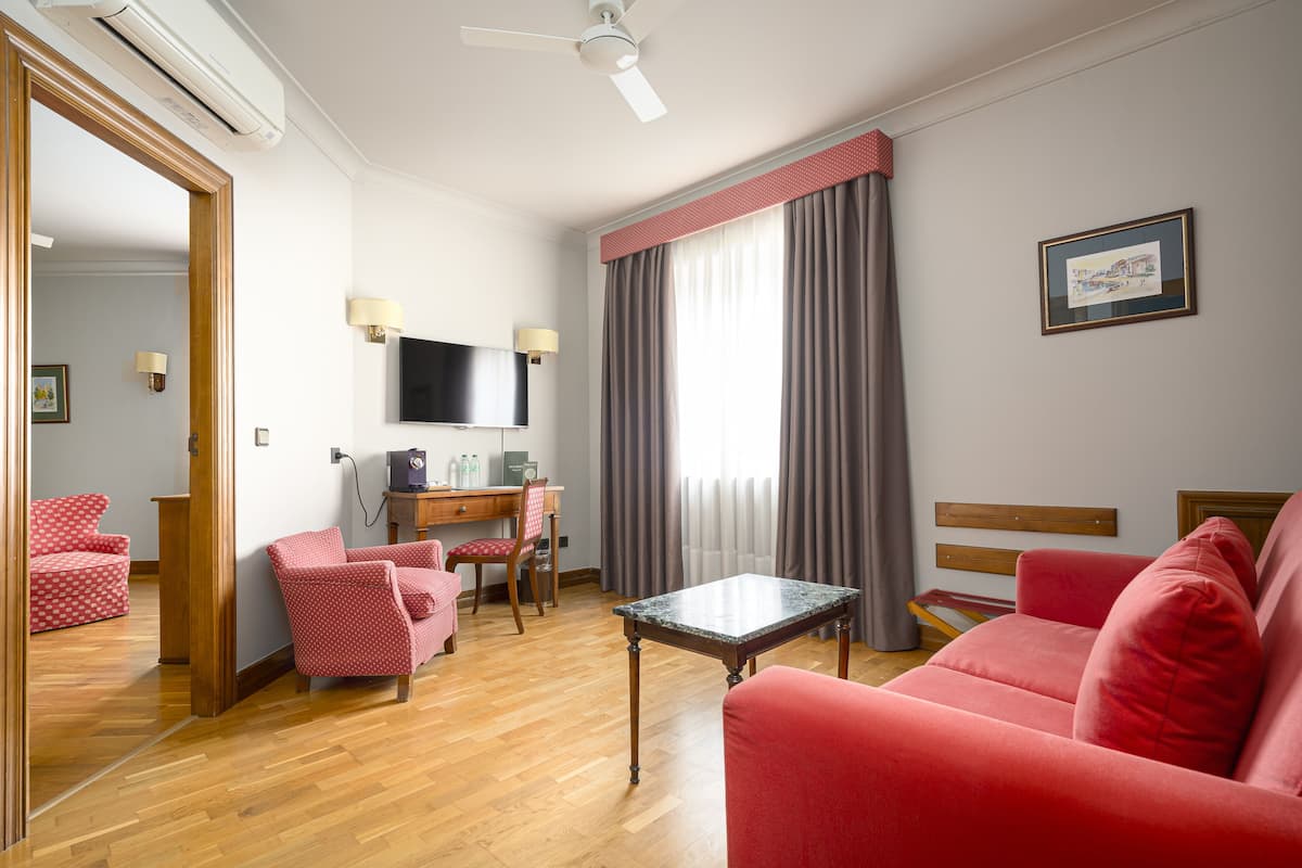 A hotel room with a wooden floor, a red sofa, a small table, a desk with a chair, a TV, and a window with curtains.