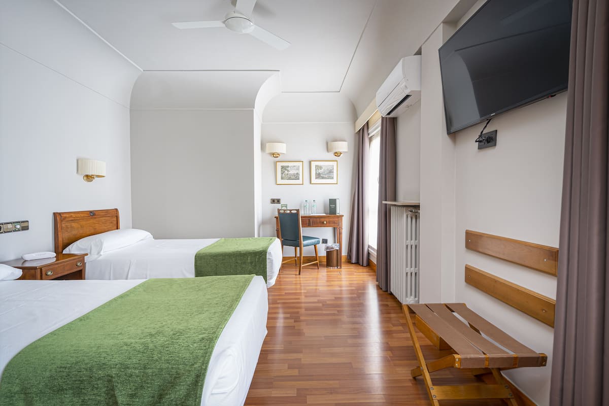 A hotel room with two beds, a desk, a chair, a TV, and a window with curtains.