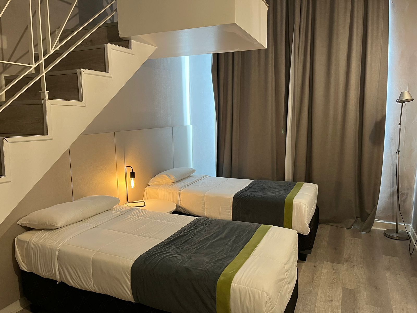 A modern hotel room with two beds, a staircase, and a large window with curtains.