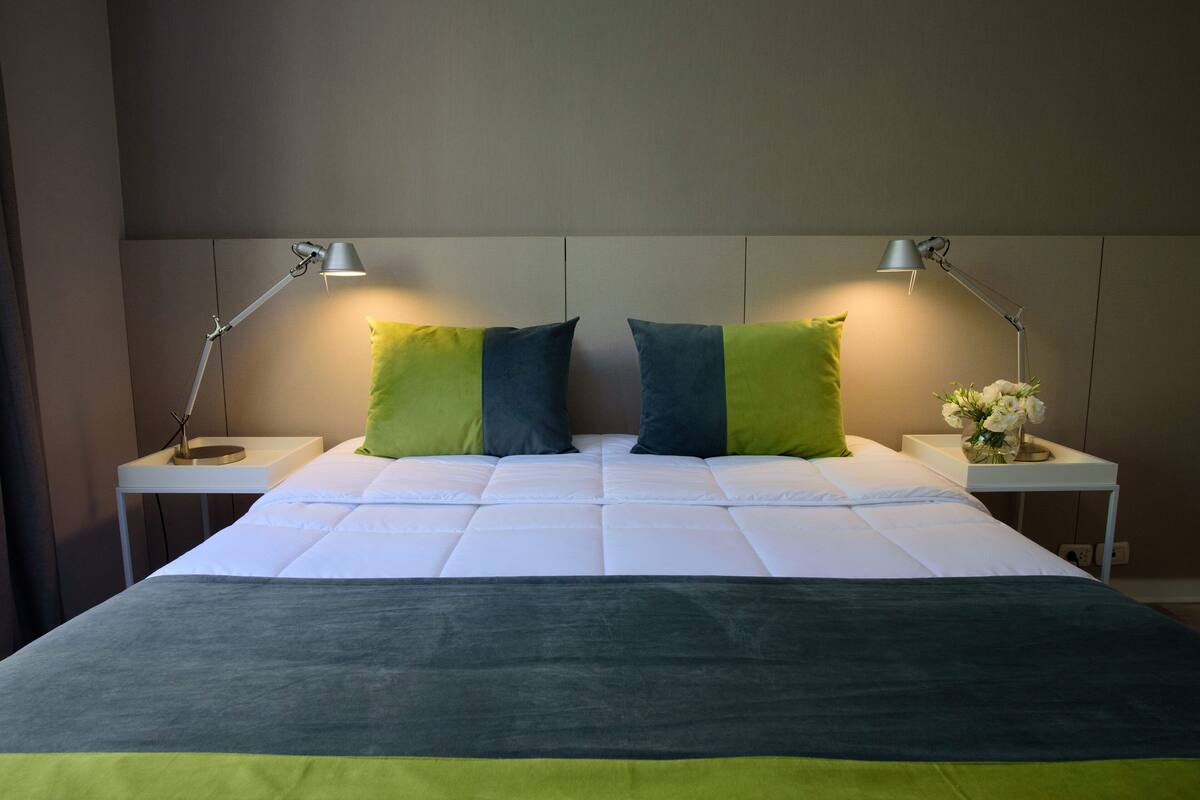A neatly made bed with a white and green bedspread, two green pillows, and two bedside lamps on white nightstands.