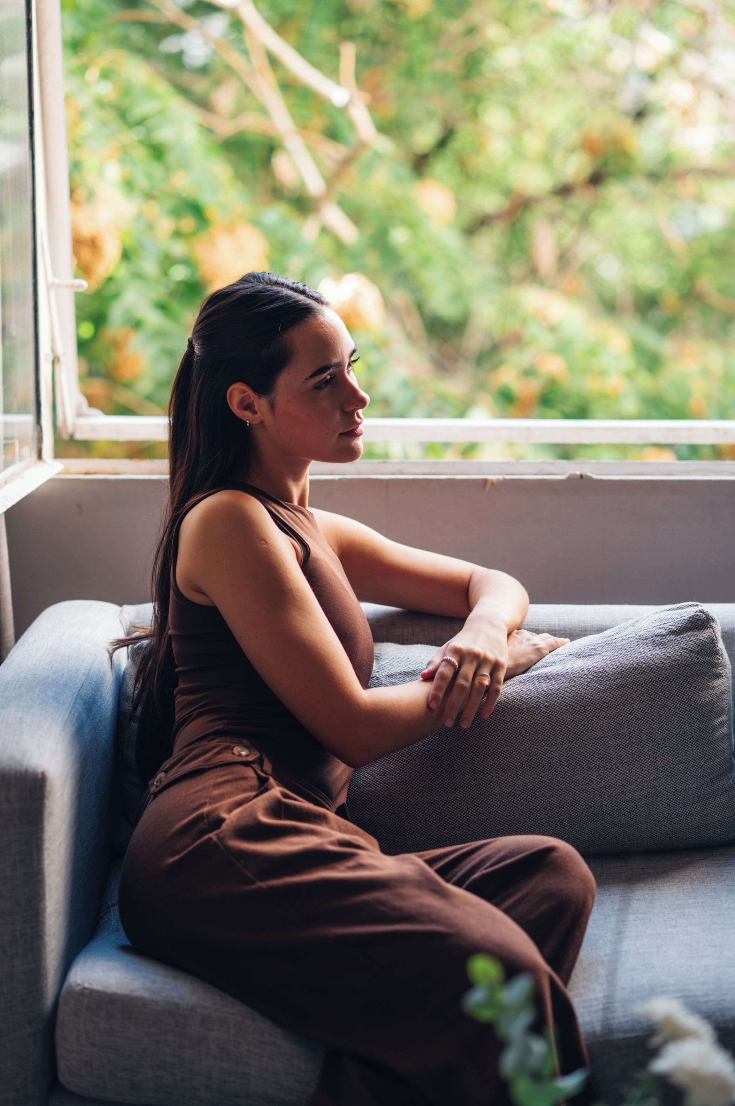 A person sitting on a grey sofa by a window with a view of greenery outside.