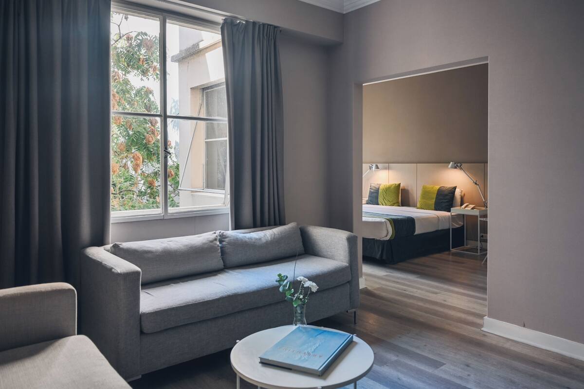 A modern living room with a sofa, a round coffee table with a book, and a view of a bedroom with a bed and bedside lamps.