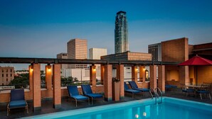 Uma piscina no terraço com espreguiçadeiras azuis e um guarda-sol vermelho, cercada por paredes de tijolo e um horizonte urbano ao fundo.