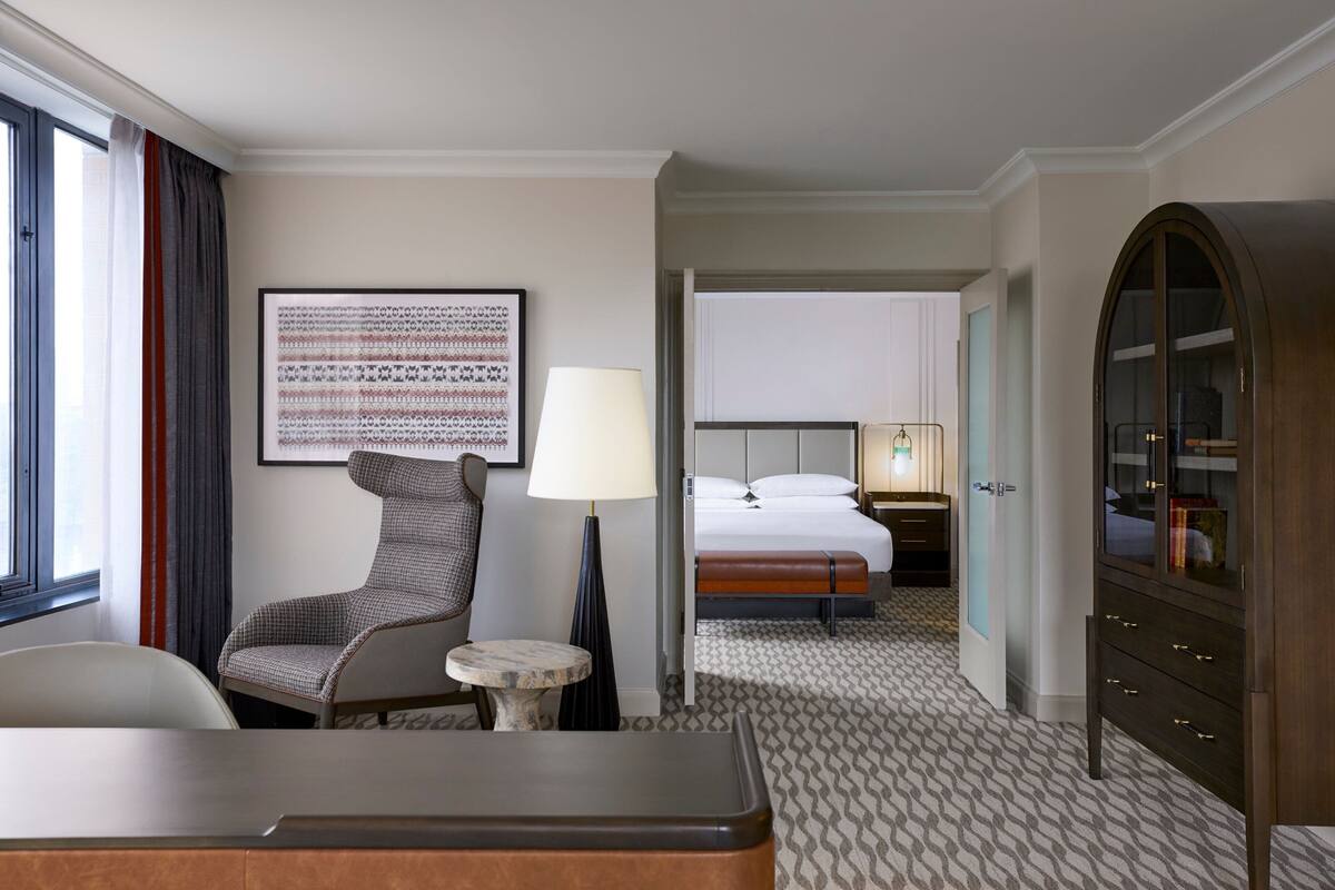 Quarto de hotel com cama, uma cadeira, uma mesa, abajur, guarda-roupa e uma obra de arte emoldurada na parede.